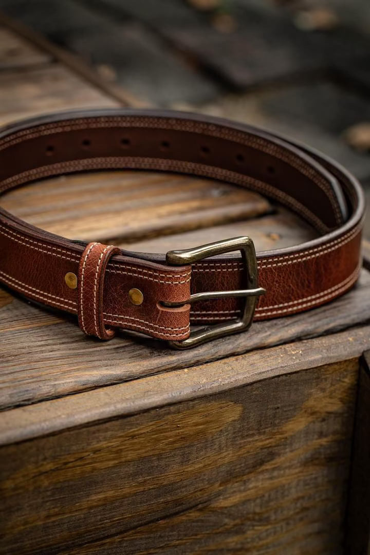 Handmade full grain leather belt in rich brown with antique brass buckle, displayed coiled on a rustic wooden surface