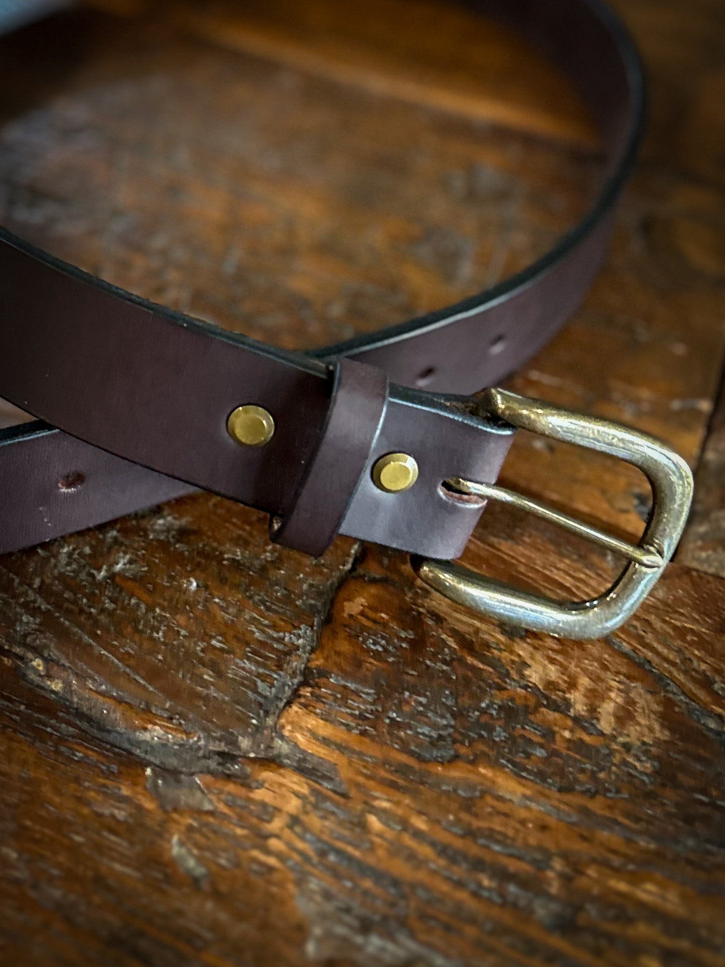 Close-up of a handcrafted brown leather belt with brass rivets and an antique brass buckle, laid on a rustic wooden surface.