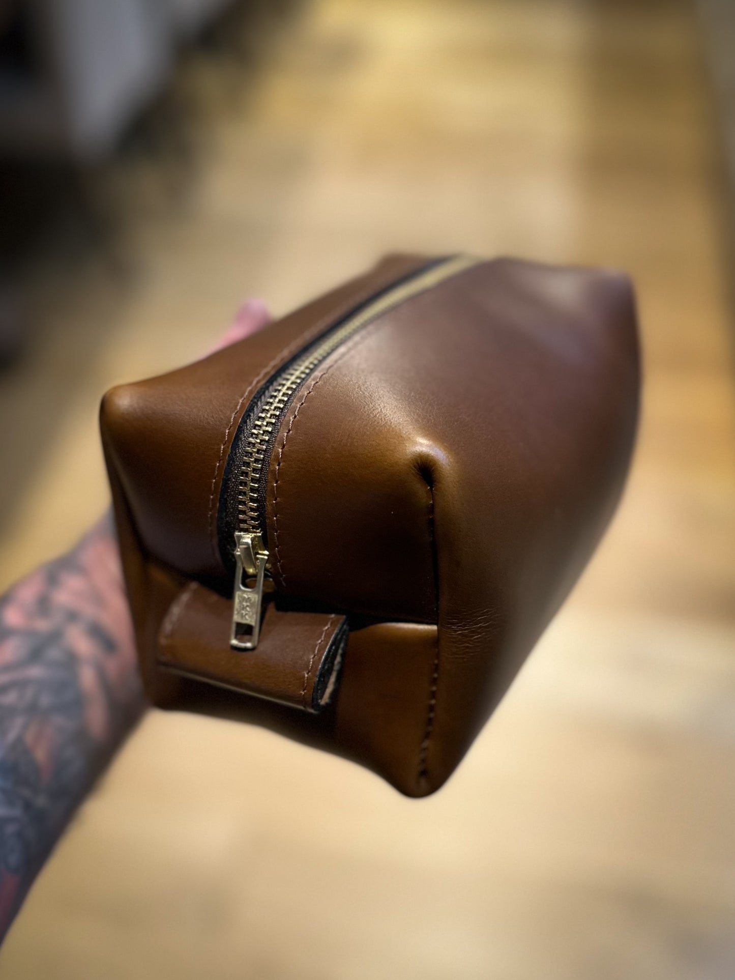 Hand holding a tan leather Dopp kit with brass zipper, showcasing the smooth full-grain leather and clean boxy construction against a warm blurred background.