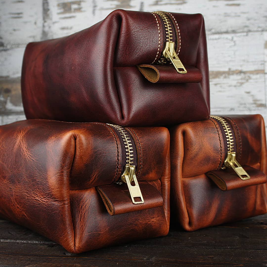 Stack of three leather Dopp kits in rich brown tones, arranged against whitewashed wooden background.
