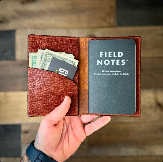 Open leather Field Notes cover in reddish-brown with notebook and cash inside, held in hand over wood background.