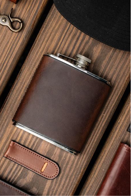 Leather-wrapped stainless steel flask on wood background with rugged accessories - handcrafted by Gunnar & CO.