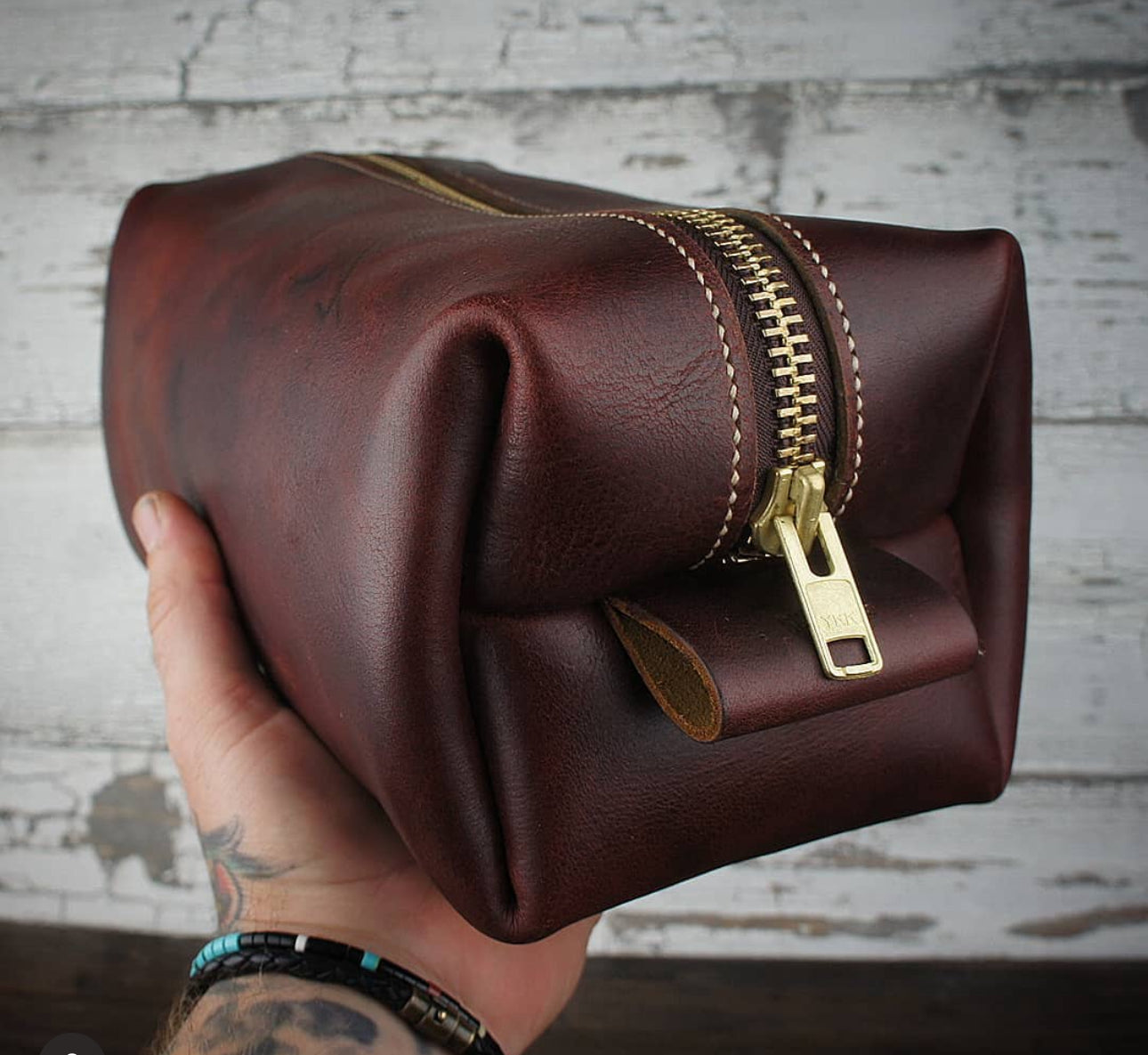 Hand holding a burgundy leather Dopp kit with a brass zipper, highlighting the curved construction.