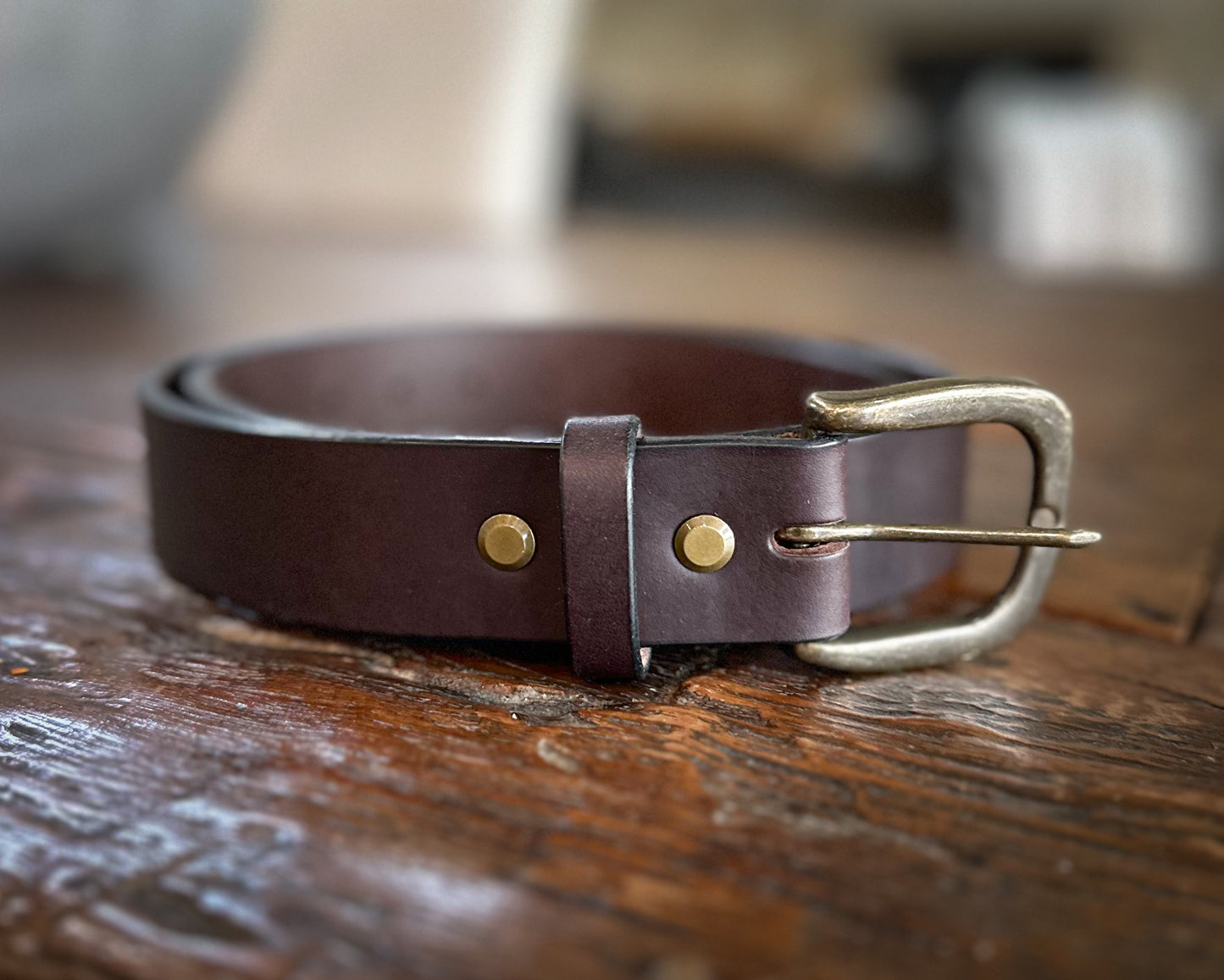 Rolled dark brown bridle leather belt featuring an antique brass buckle and double brass rivets, displayed on a rustic wooden surface.