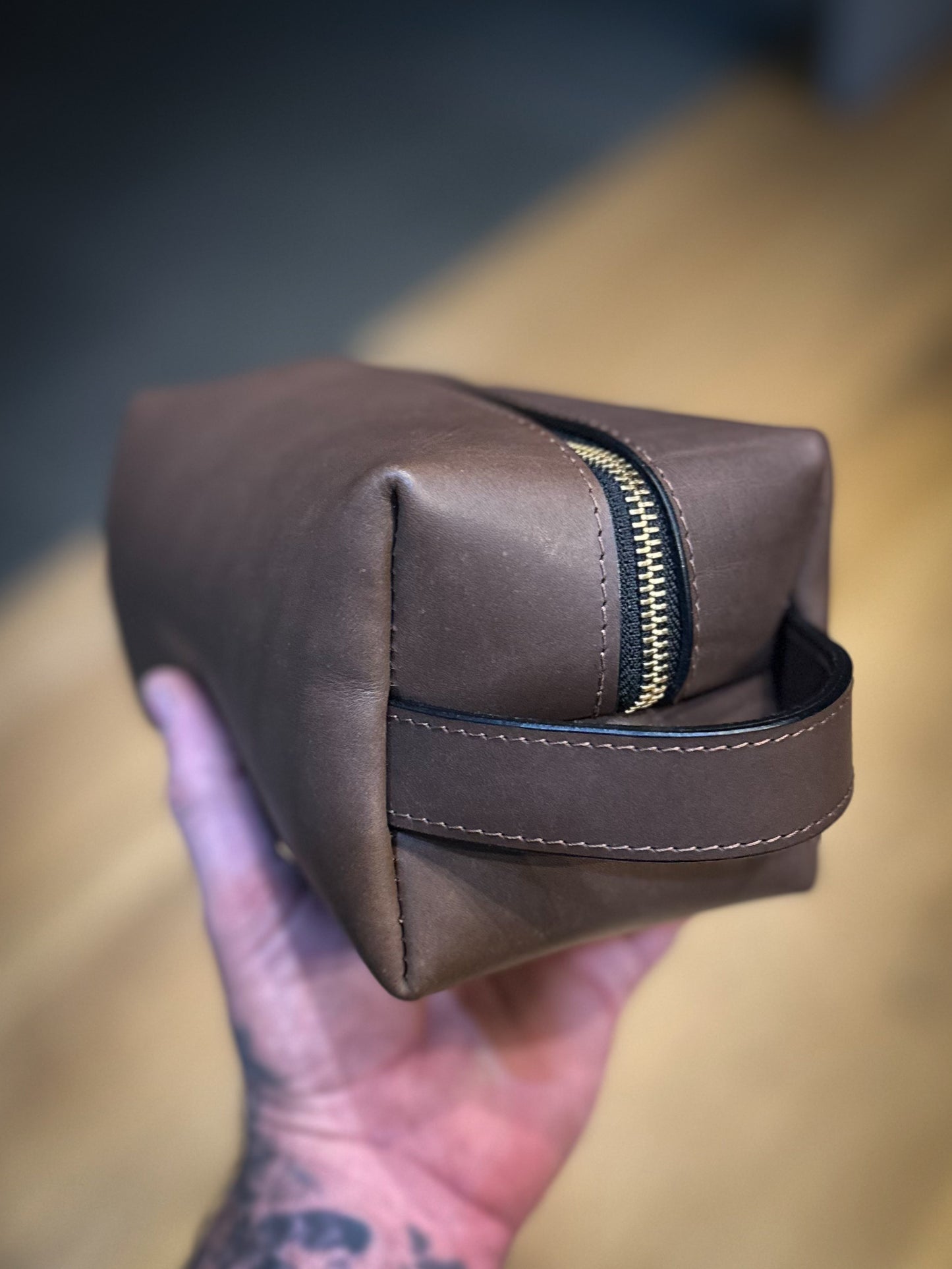 Smooth chocolate brown leather Dopp kit with loop handle and contrast stitching, held in hand.