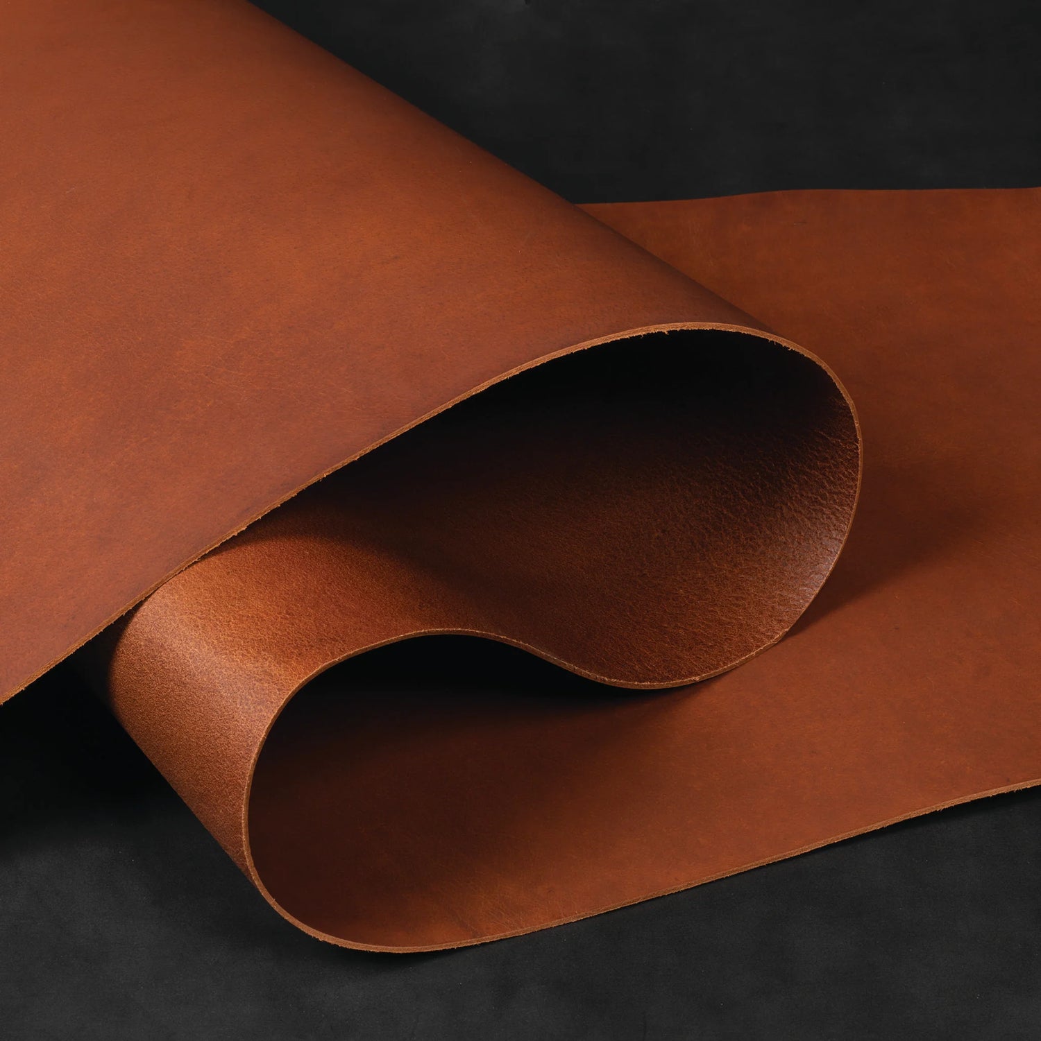 Close-up of a rolled side of full-grain leather in rich brown tones, showing natural texture and grain on a black background.