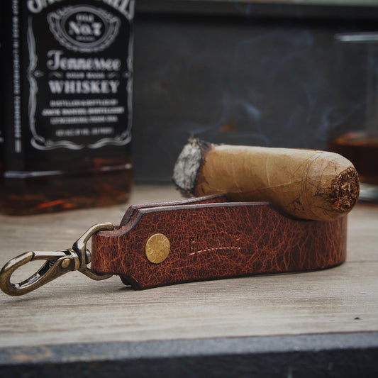 Leather cigar rest keychain beside a cigar and whiskey bottle on wooden surface