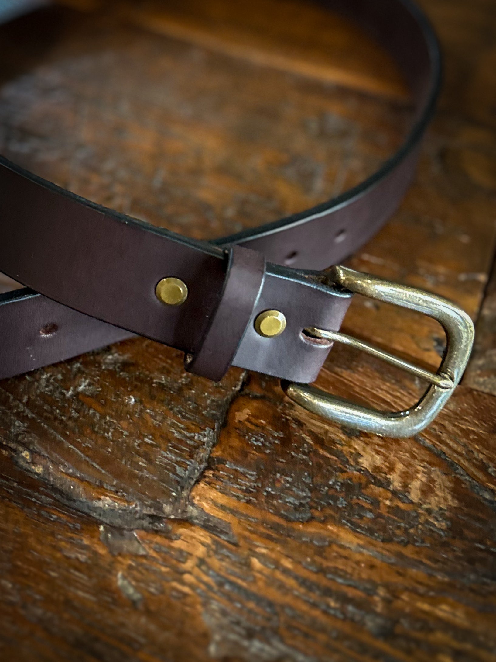 Close-up of a handcrafted brown leather belt with brass rivets and an antique brass buckle, laid on a rustic wooden surface.