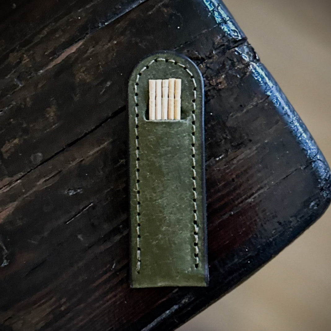 Handcrafted leather toothpick holder in green, shown on a rustic wooden background. Features white stitching and a visible row of toothpicks.
