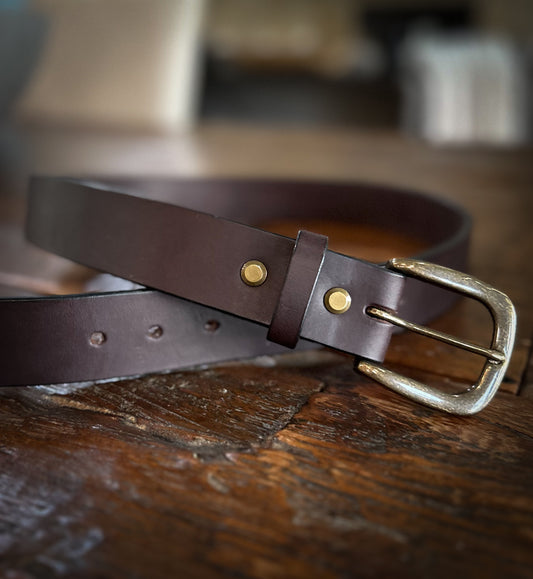 Full-grain bridle leather belt with antique brass buckle and double brass rivets, laid flat and coiled on a rustic wood surface.