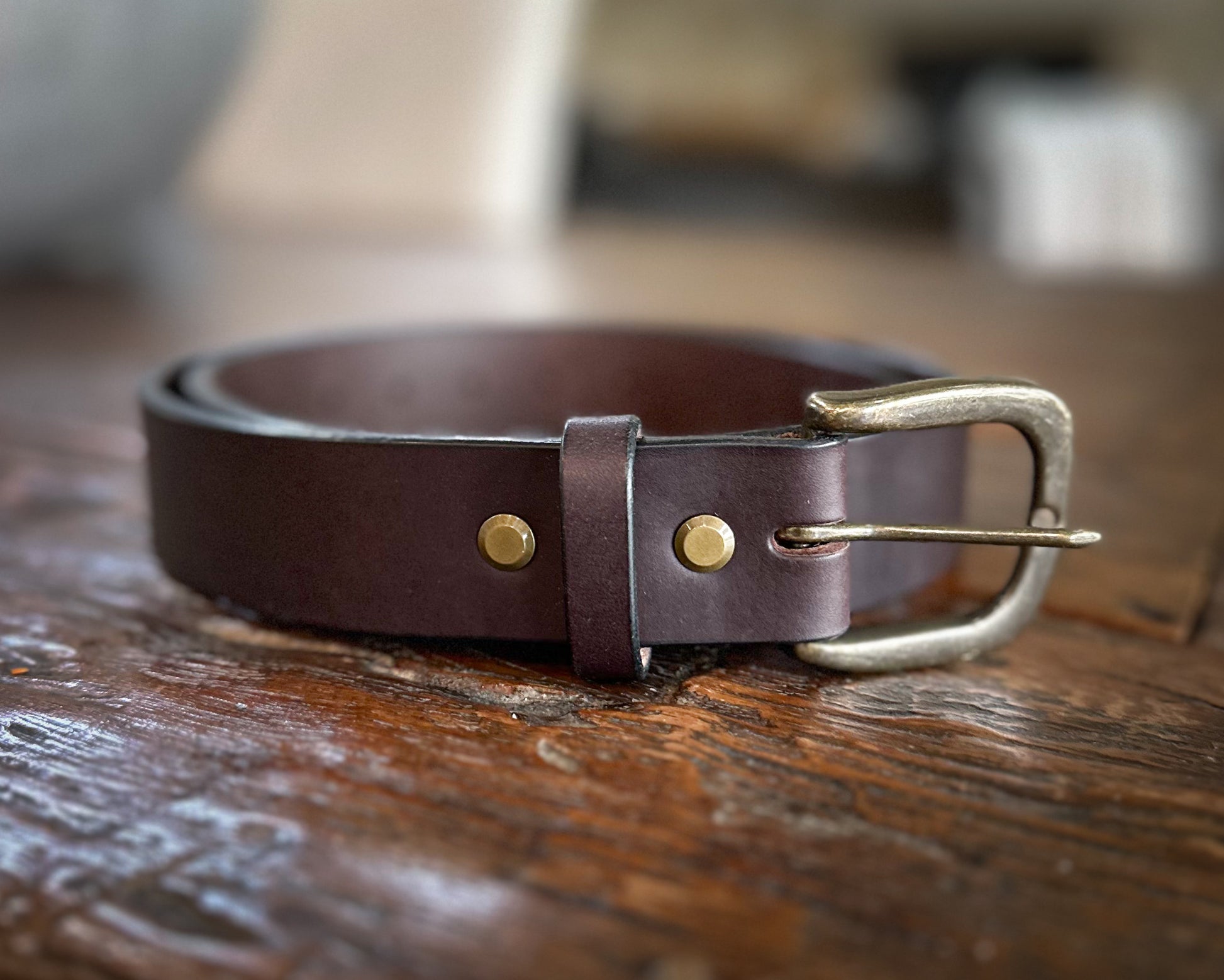 Rolled dark brown bridle leather belt featuring an antique brass buckle and double brass rivets, displayed on a rustic wooden surface.