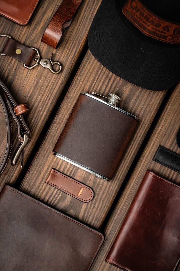 Leather-wrapped flask styled with handmade leather goods and Gunnar & CO hat - flat lay on rustic wood background