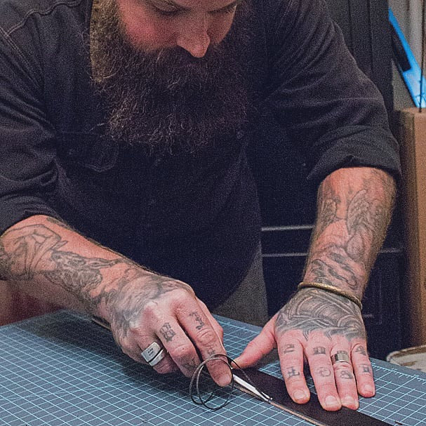 Close-up of a leather craftsman cutting leather by hand with a round knife on a cutting mat. Tattooed forearms and focused detail work reflect hands-on craftsmanship.