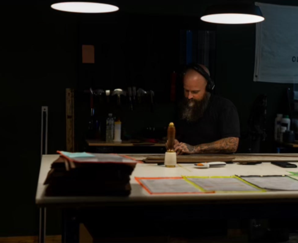 Brandon Pangle of Gunnar & Co. working at a leathercraft bench under warm lights, reviewing production materials in a workshop setting.